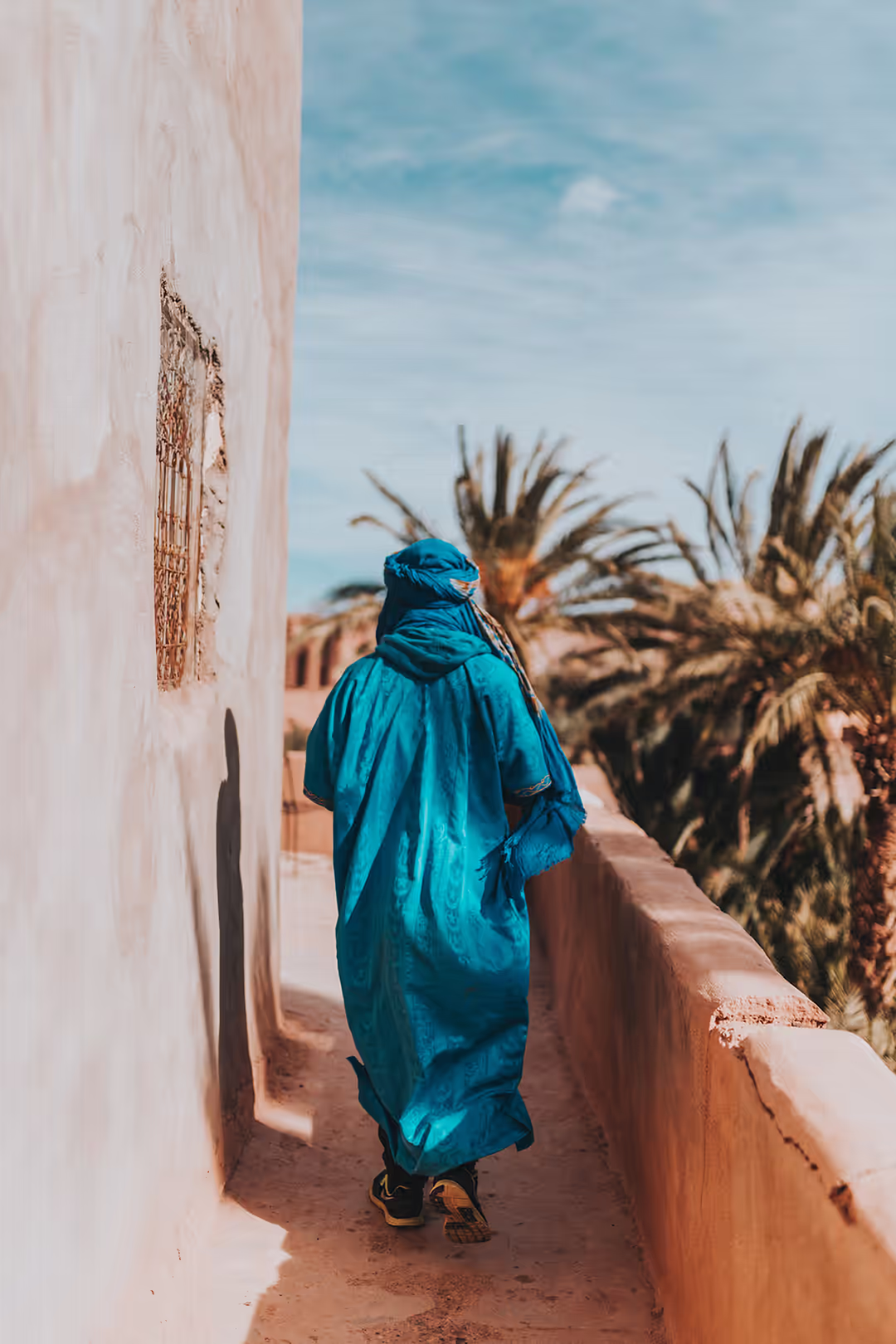 Hombre con gandora azul caminando junto a una muralla y palmeras